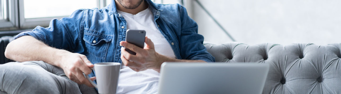 A man holding a cup of coffee while holding his cell phone and balancing a laptop on his lap.